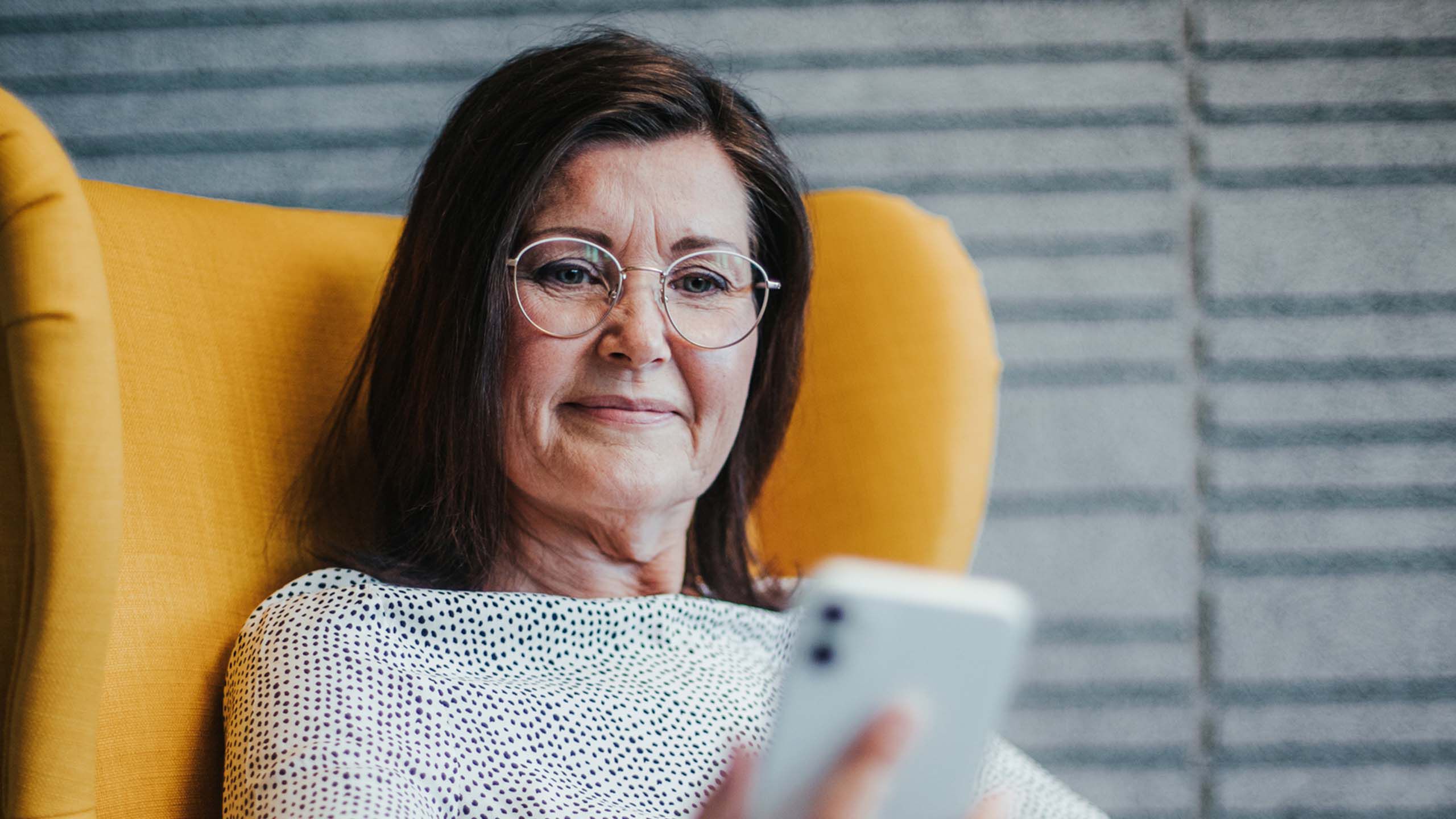 Ik&auml;&auml;ntynyt nainen istuu silm&auml;lasit p&auml;&auml;ss&auml; nojatuolissa ja katsoo puhelinta.