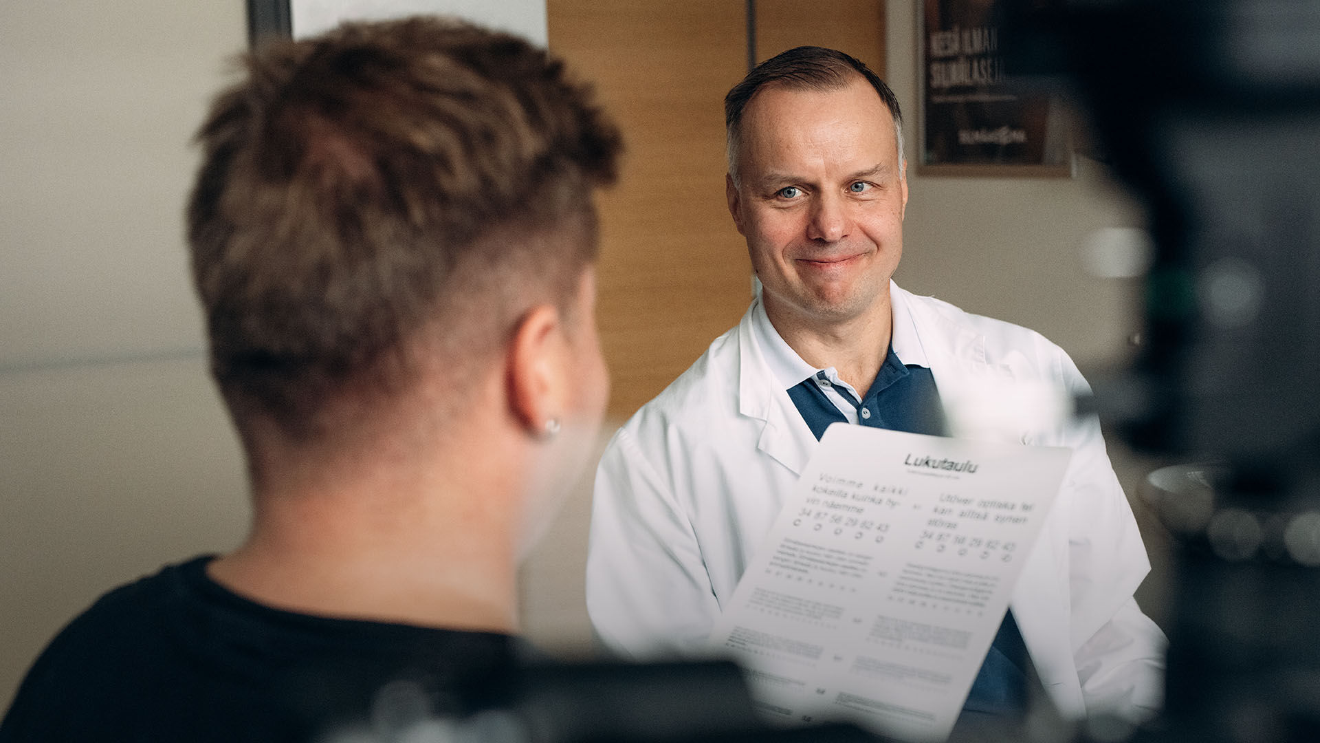 Vastaanottohuoneessa silm&auml;l&auml;&auml;k&auml;ri ohjeistaa asiakasta lukemaan teksti&auml; n&auml;&ouml;ntarkastusta varten