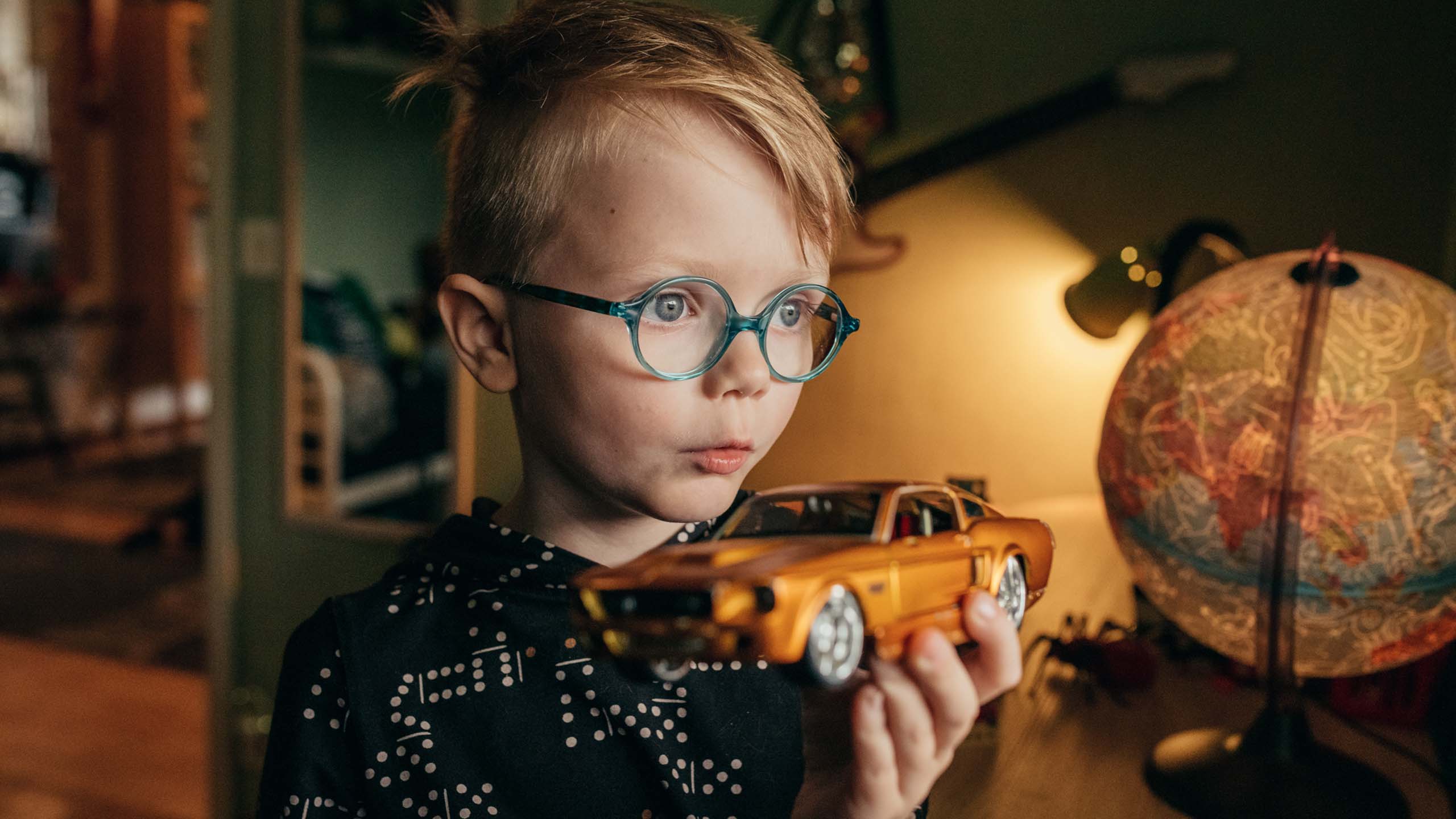 Silmälasipäinen lapsi sisätiloissa auto kädessä