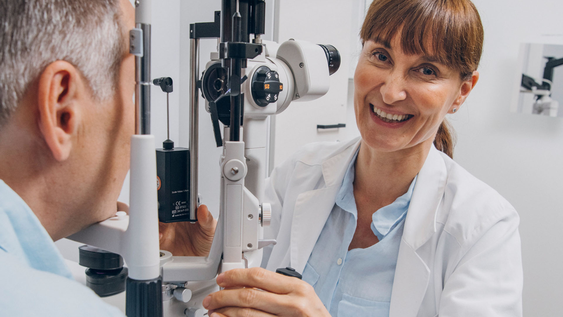 Silmälääkäri hymyilee kameralle vastaanottohuoneessa mikroskoopin takaa