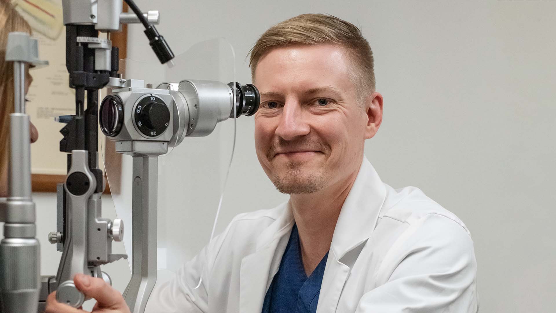 Silmälääkäri hymyilee tutkimuslaitteen takana kameralle