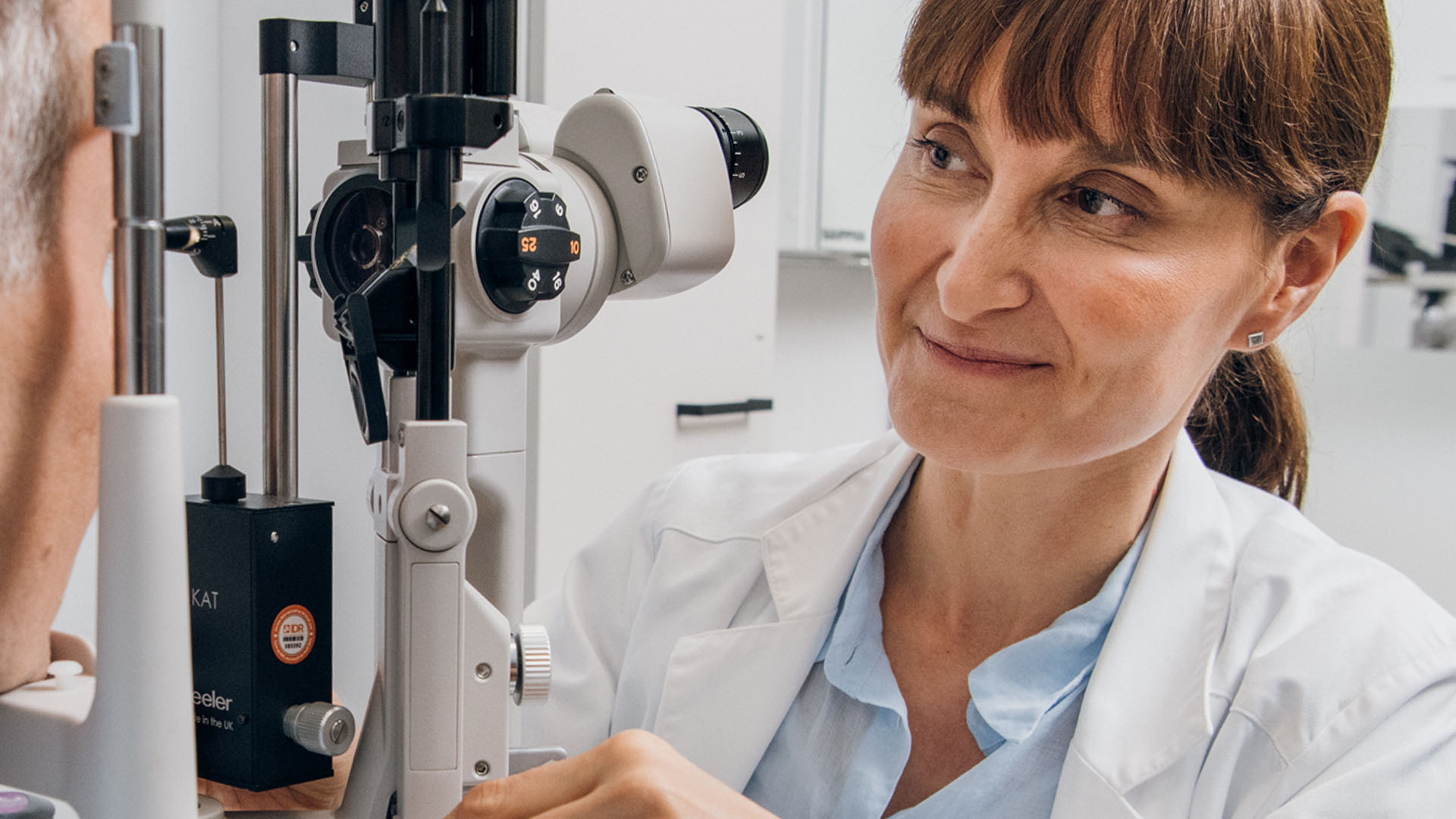 Ystävällinen silmälääkärinainen hymyilee asiakkaalle joka istuu mikroskoopin takana