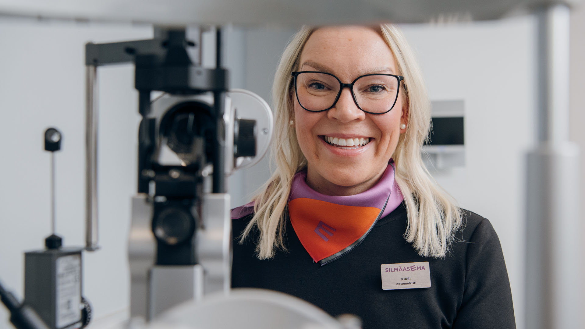 Optikko hymyilee näöntutkimuslaitteen takana kameralle