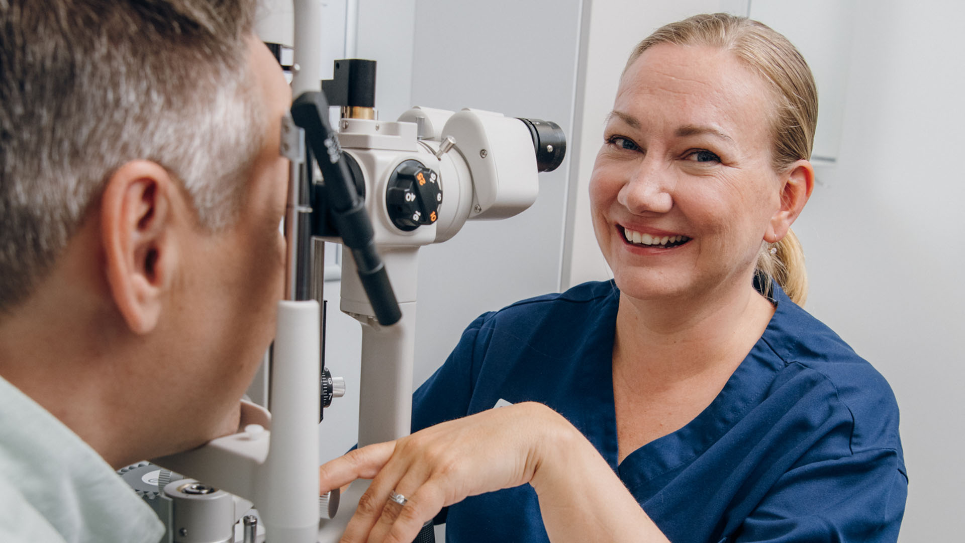 Kuivasilmäklinikan optikko hymyilee kameralle, asiakas mikroskoopin takana tutkimuksessa.