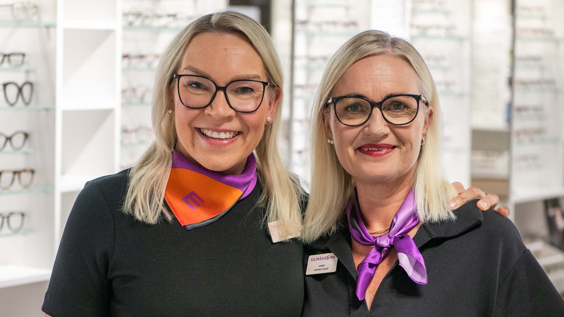 Silmäaseman optikko ja myyjä myymälässä kameralle hymyillen