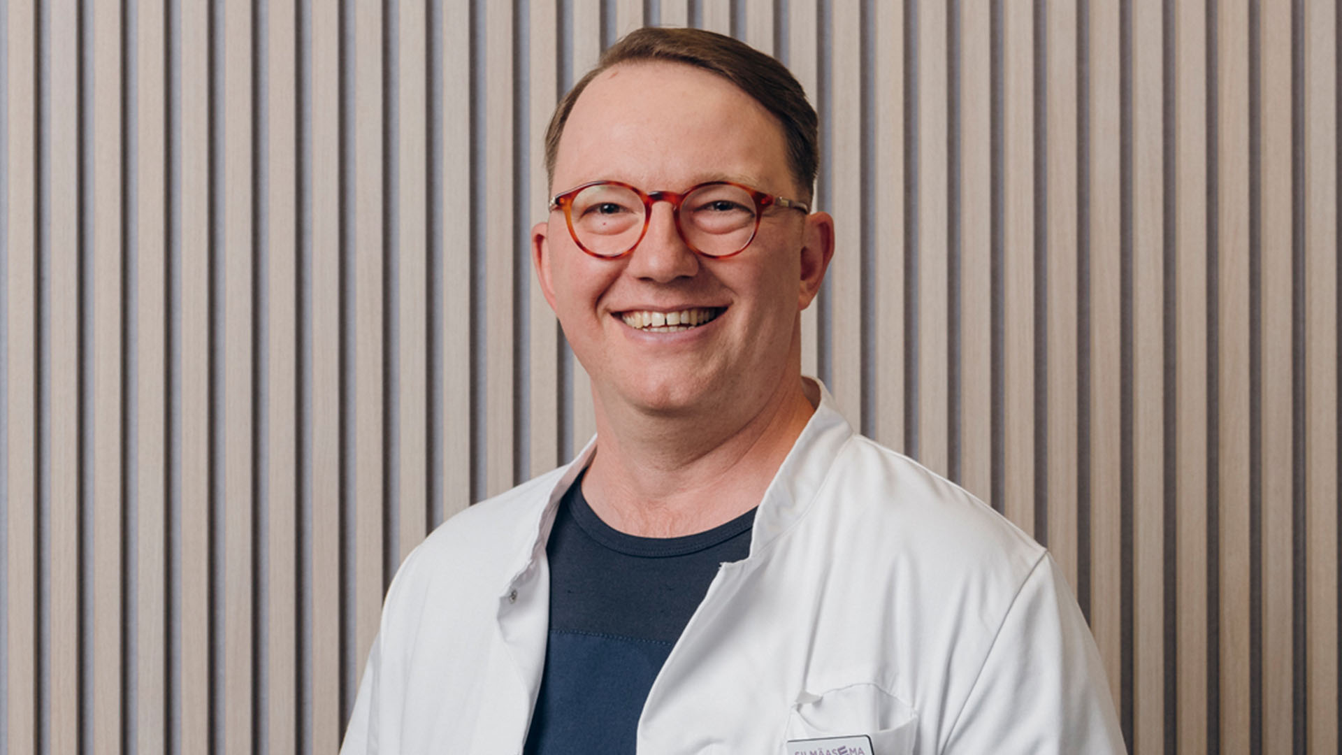 Silmälääkäri hymyilee kameralle rimaseinän edessä