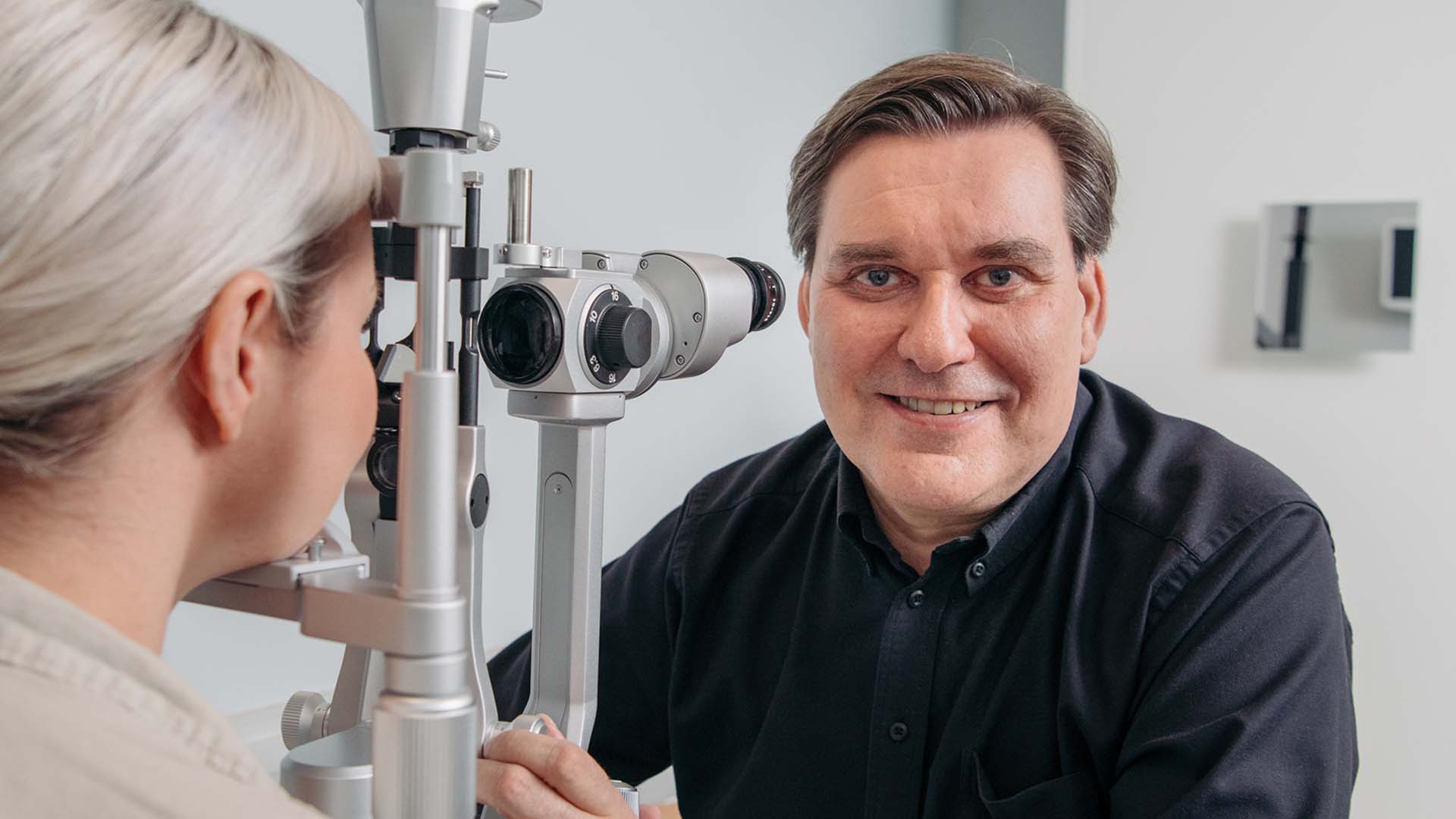 Optikko hymyilee kameralla näöntutkimuslaitteen takana
