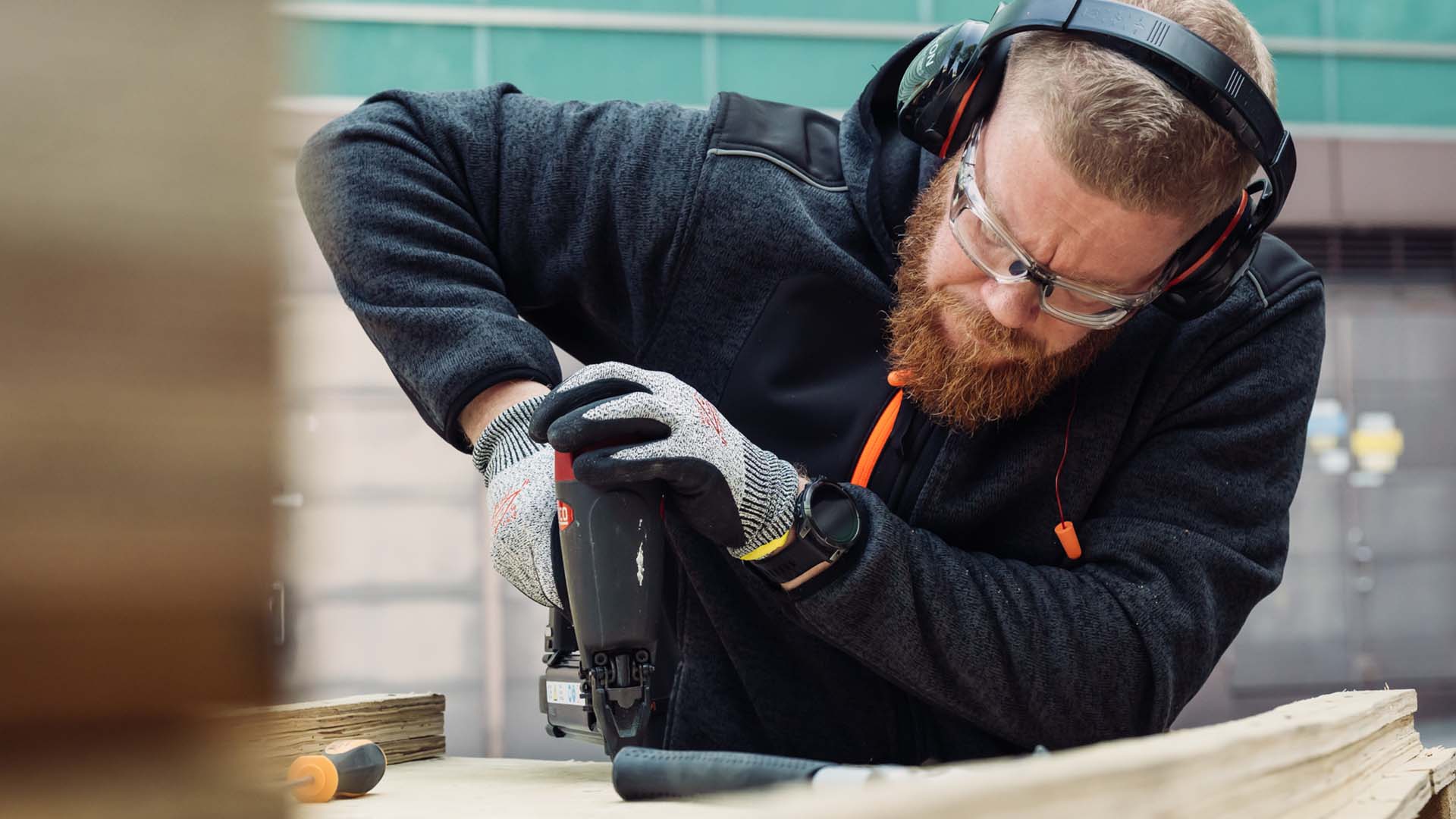 Suojalasit ja kuulosuojaimet päässä työskentelevä parrakas mies.