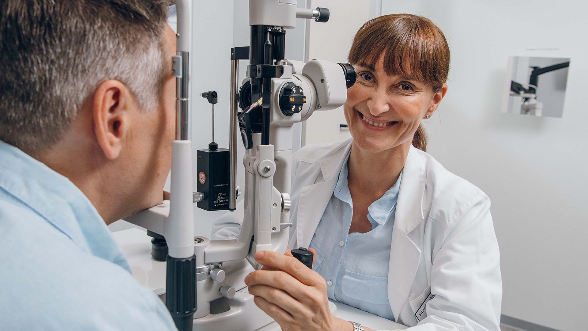 An ophthalmologist smiles at the camera from behind a microscope in an examination room.