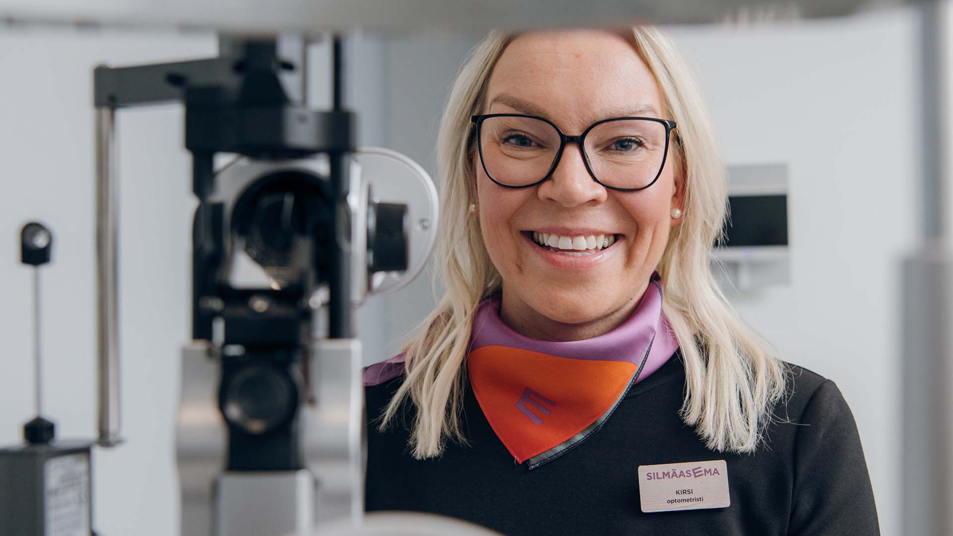 Iloisesti hymyilevä optikko näöntutkimuslaitteen takana