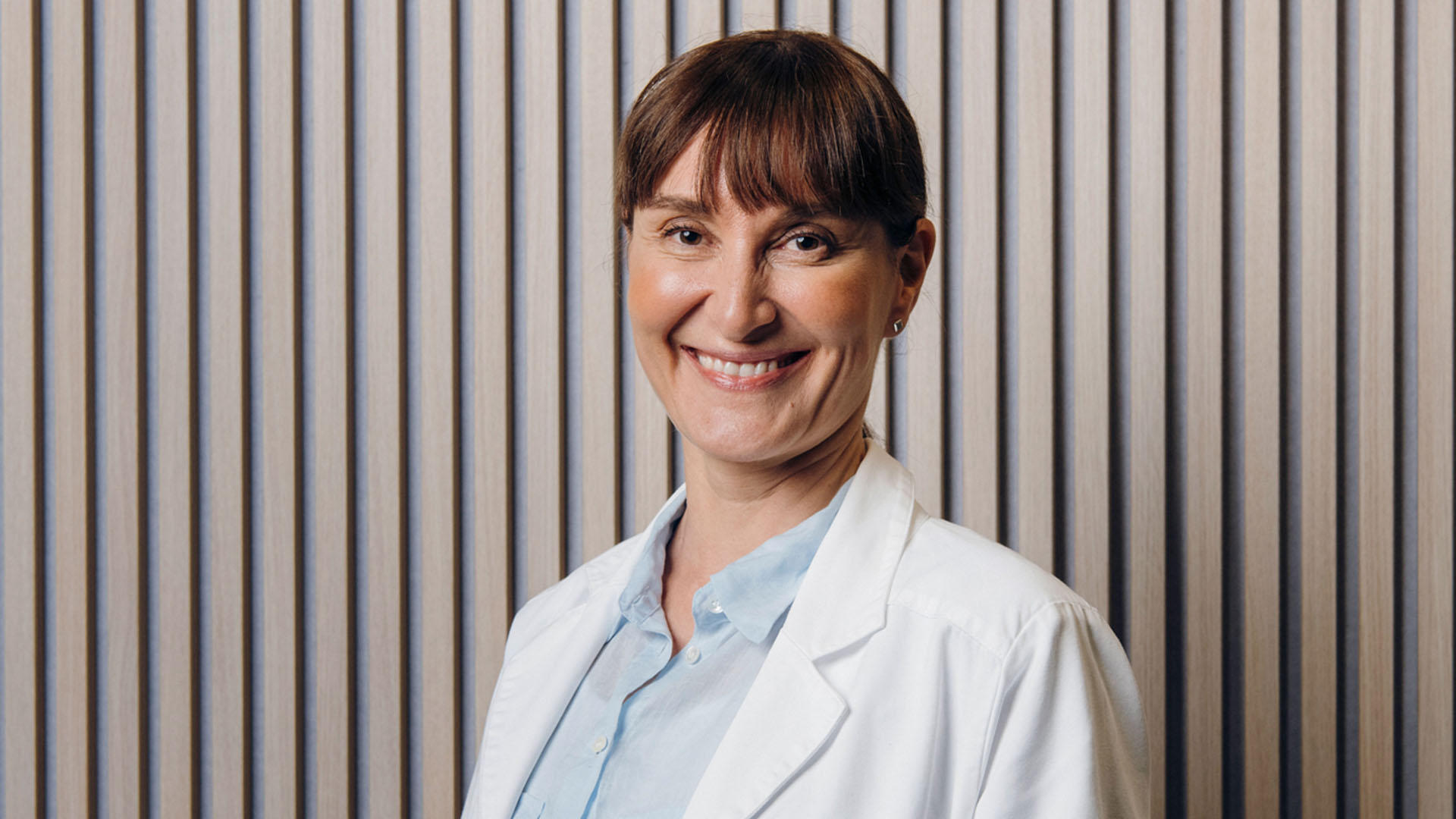 Naispuolinen silmälääkäri hymyilee kameralle rimaseinän edessä