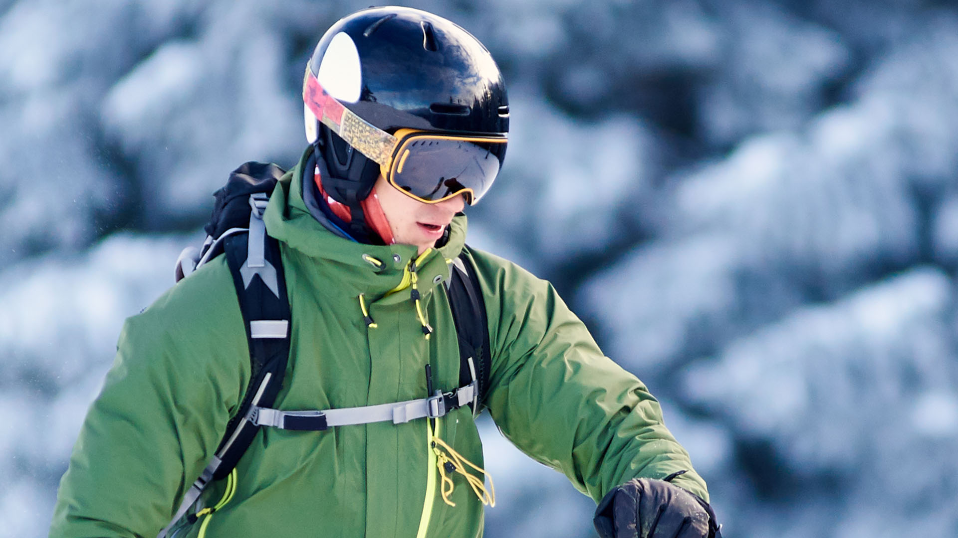 Laskettelulasit kasvoilla oleva lumilautailija, mies vihreässä takissa ja reppu selässä.