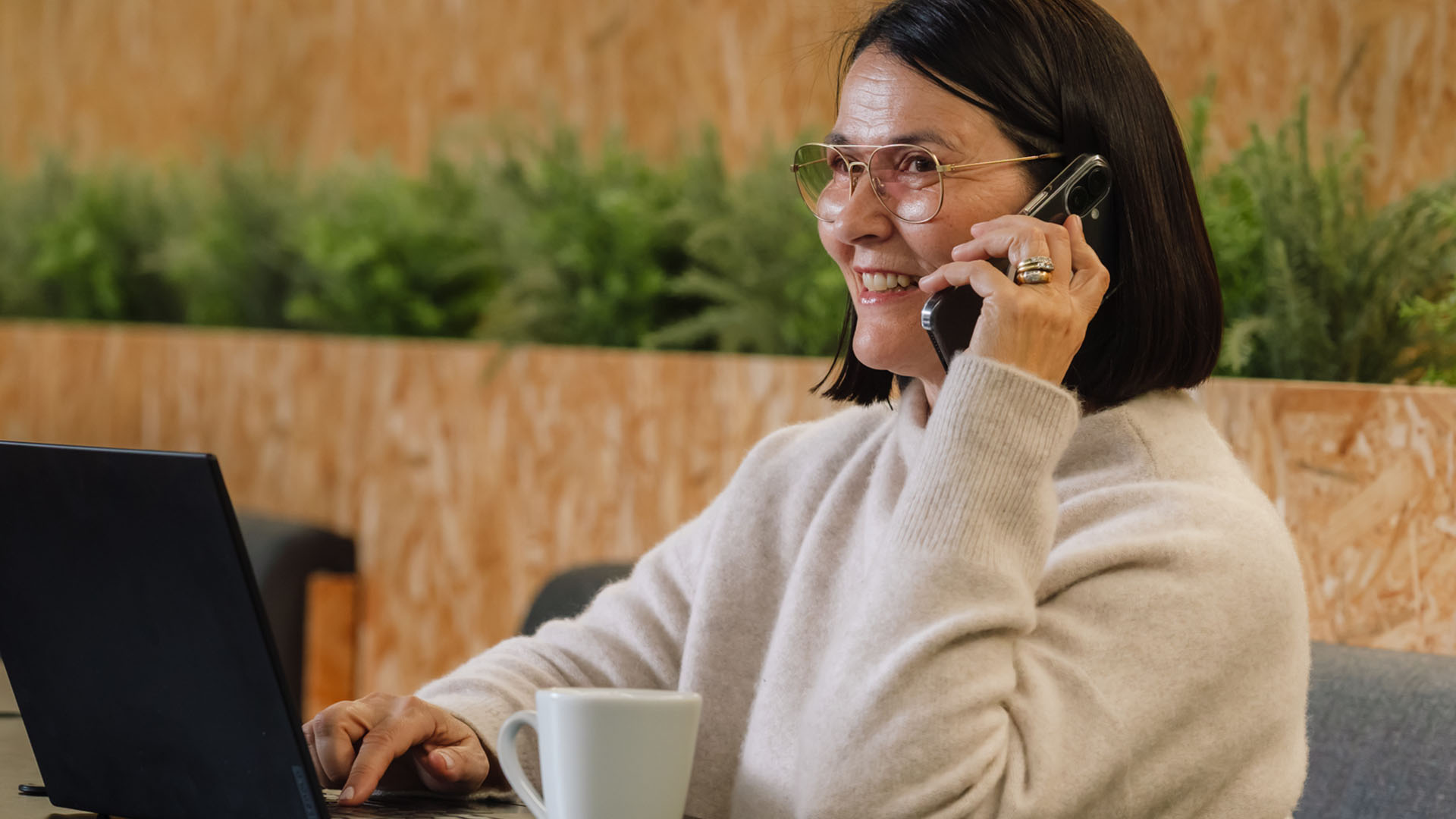 Etänä ergonomisesti työskentelevä nainen läppäri pöydällä ja puhelin korvalla.