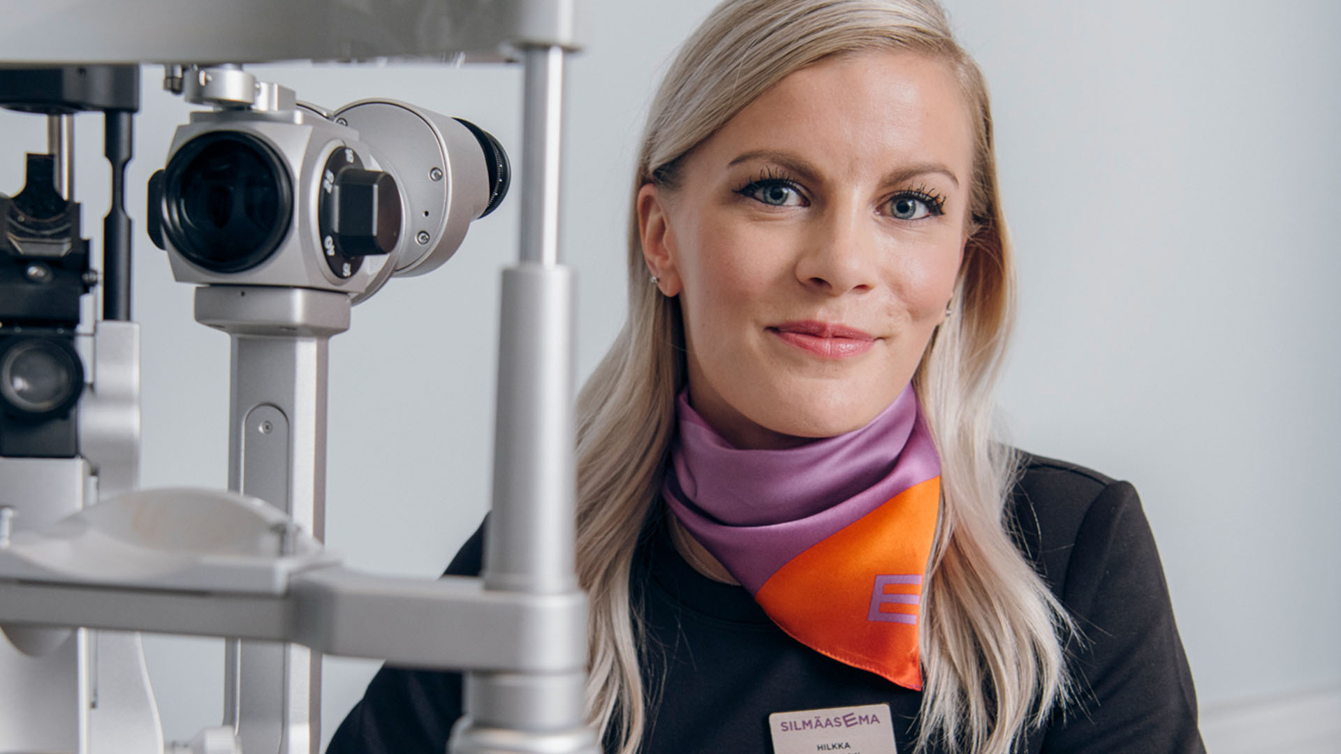 Optikko näöntutkimuslaitteen takana kameralle hymyillen