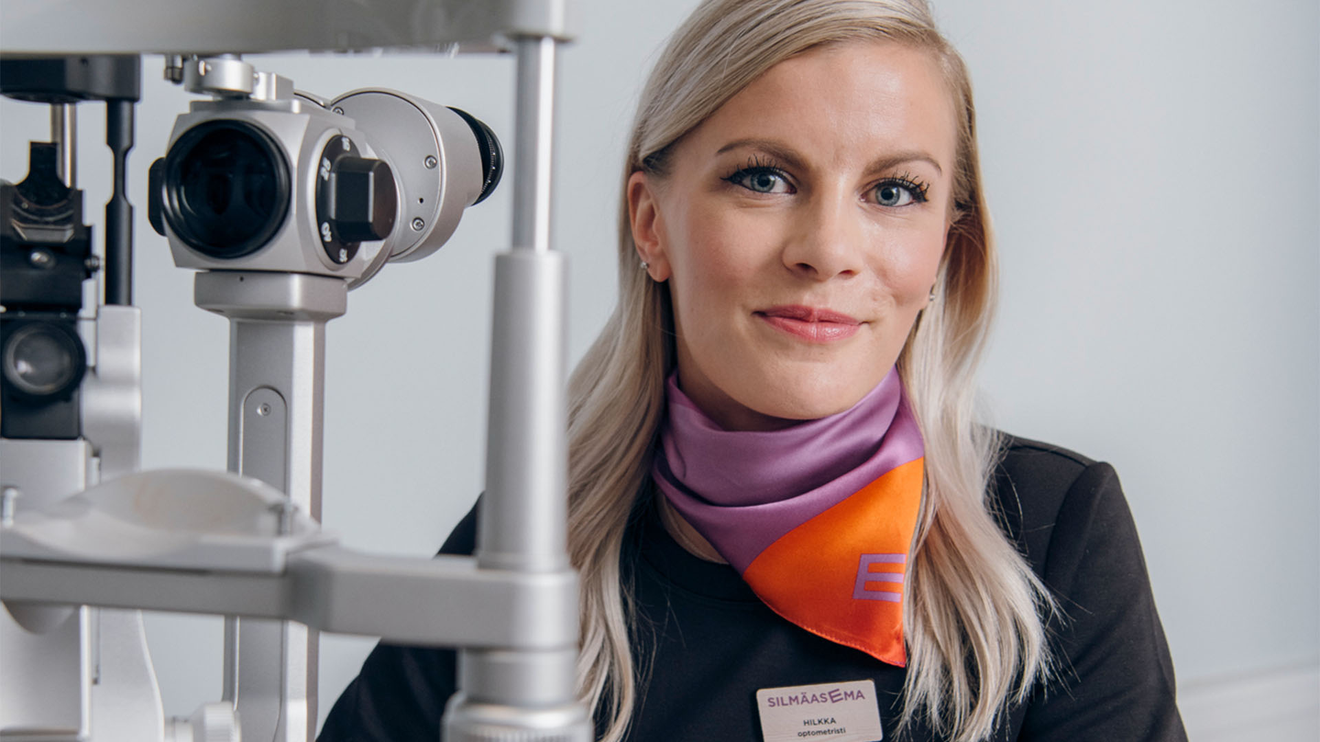 Optikko hymyilee kameralle näöntutkimuslaitteen takaa.