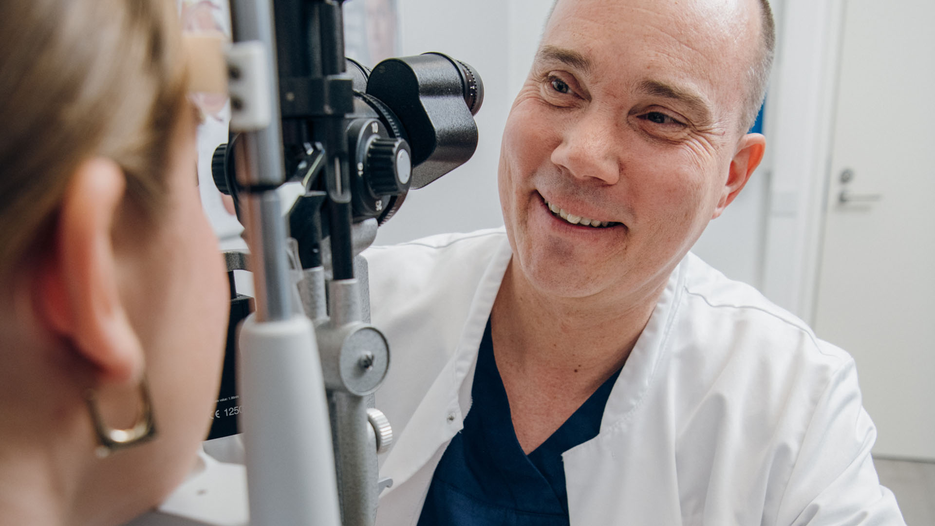 Ystävällinen silmälääkäri hymyilee asiakkaalle joka istuu mikroskoopin toisella puolella