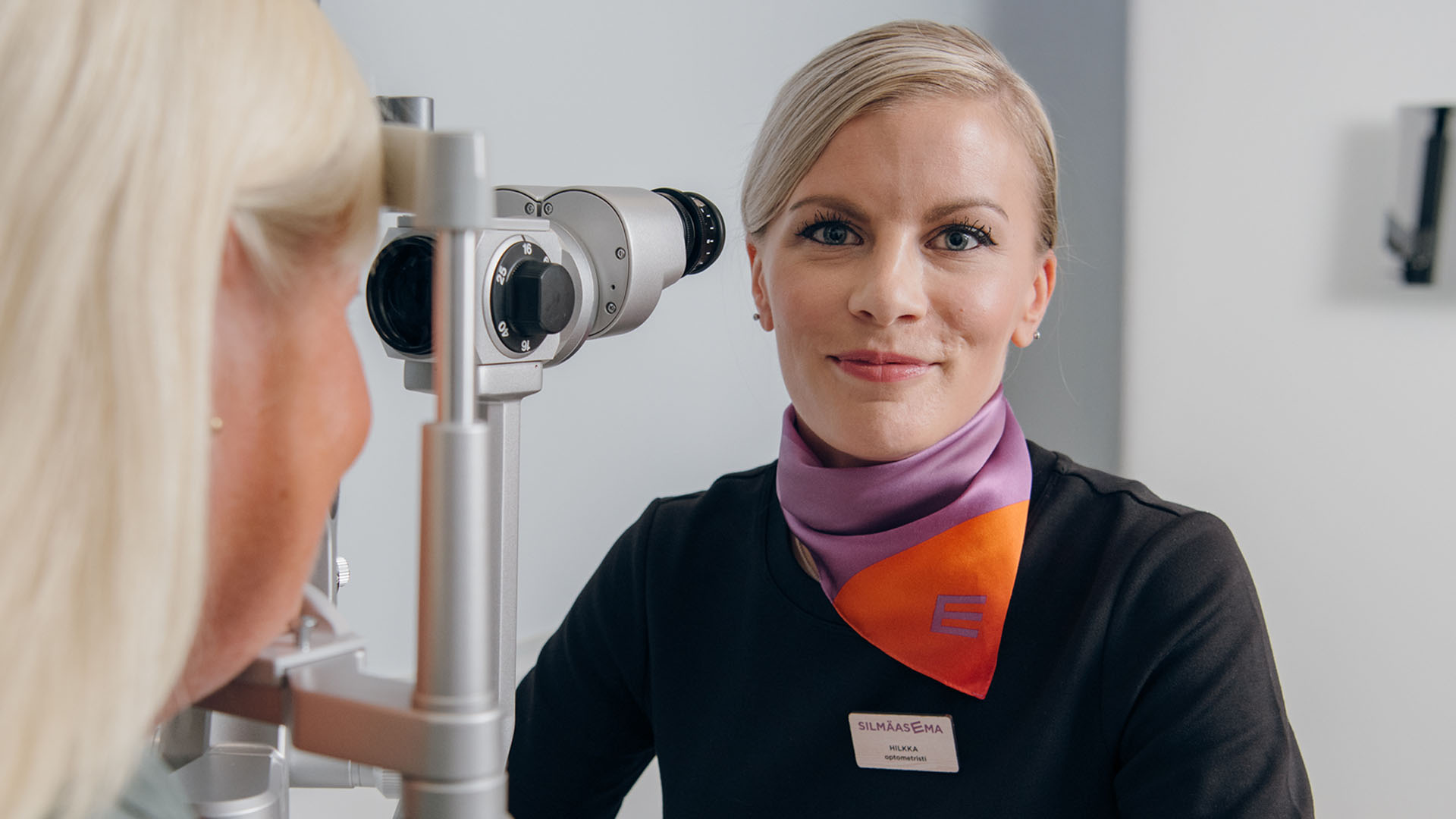 Optikko näöntutkimuslaitteen takana kameralle hymyillen.