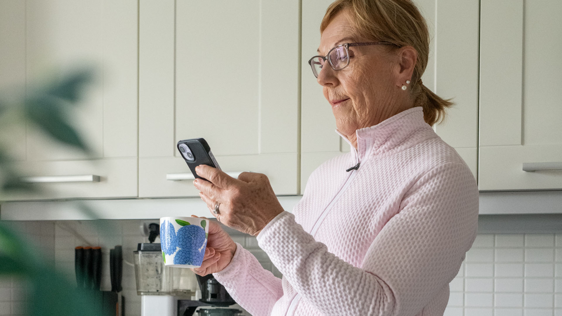Kaihi-ikäinen rouva seisoo keittiössä kahvikuppi kädessä puhelinta katsoen
